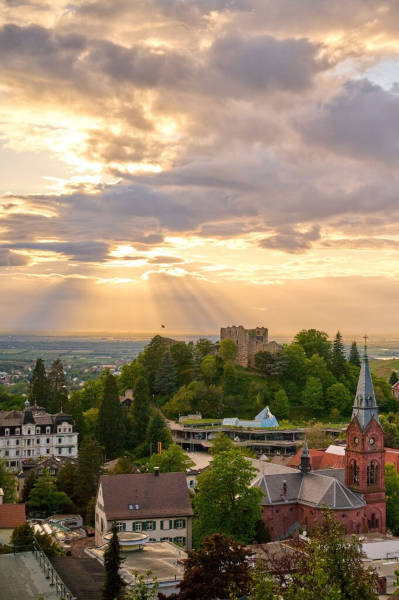 Die Abendsonne streckt ihre Strahlen durch die Wolkendecke. Auf der unteren Hälfte des Bilds ist ein städtisches Viertel mit vielen Bäumen und einem kleinen roten Schloss.