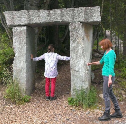 Frau steht in einem Steintor, eine weitere Frau steht mit einem Pendel in der Hand daneben.