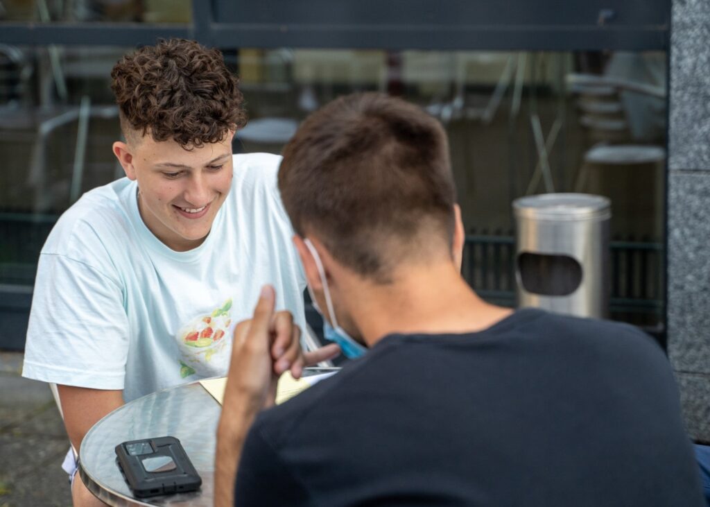 Zwei junge Männer sitzen an einem Tisch. Derjenige, der zur Kamera schaut lächelt, vom anderen ist nur der Hinterkopf abgebildet. Dieser hält sich die Hand in der Telefon-Geste ans Ohr.