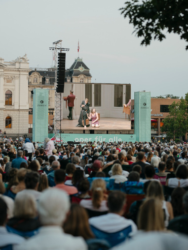 Eine Menschenmenge blickt zur Leinwand, auf der eine Oper gezeigt wird.