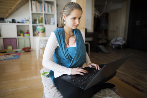 Junge Frau zuhause am Laptop, Baby vor der Brust in Tragetuch