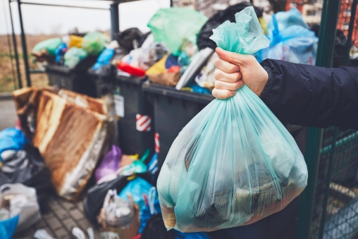 Eine Hand entsorgt eine Mülltüte, im Hintergrund stehen volle Müllcontainer Better Recycling