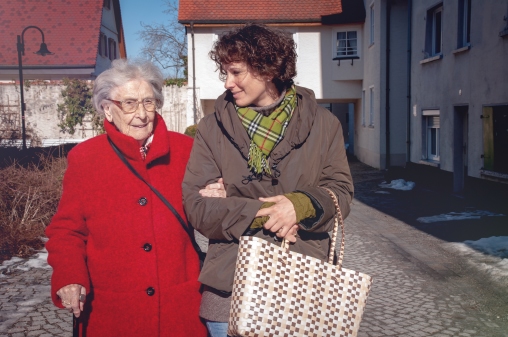 Eine Frau begleitet eine ältere Dame auf einem Spaziergang, den Arm eingehakt